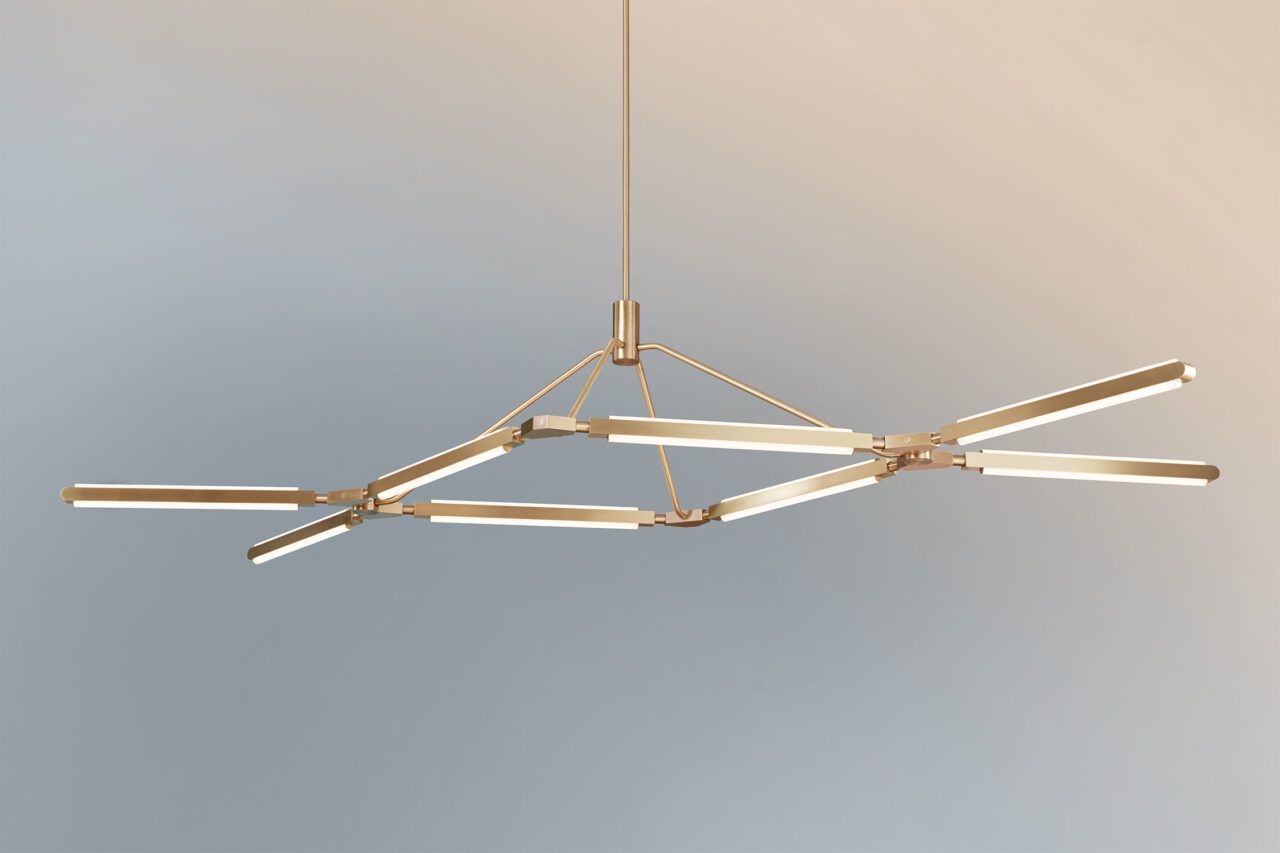 Three quarter view of an illuminated Pris Crown chandelier with satin brass metal components in front of a peach to dusty blue gradient.