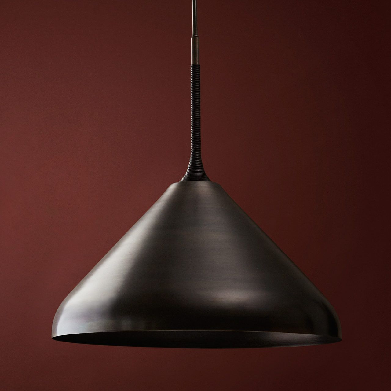 Pelle’s Stem Shade 20 in black brass with black leather coiling hangs against a deep red background.