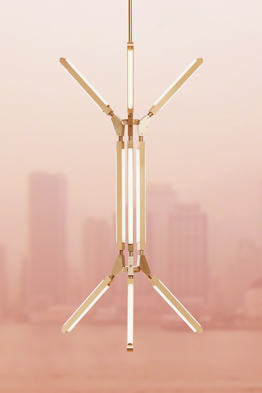Eye level view of an illuminated Pris Blaze chandelier with satin brass metal components in front of a foggy peach-toned cityscape.